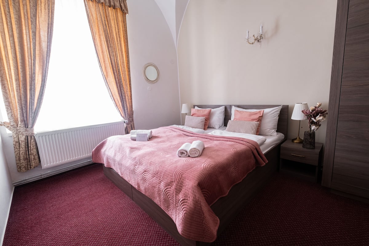 A neatly made bed with a pink blanket and pillows, a bedside table with a lamp and a vase, and a window with curtains.