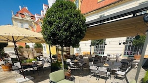 Un área de estar al aire libre con mesas y sillas, un árbol pequeño y un edificio al fondo.