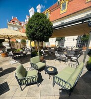 An outdoor seating area with tables and chairs, a small tree, and a building in the background.