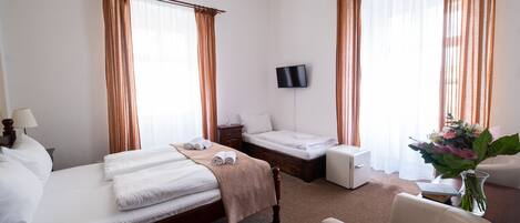 Habitación de hotel con cama, mesita de noche, televisión, silla y escritorio con un jarrón de flores.