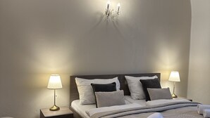 Una cama bien hecha con un edredón gris, dos lámparas de noche y una mesita auxiliar pequeña.
