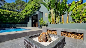 Un espace piscine extérieur moderne, doté d’un foyer et d’une pile de bois, entouré d’une végétation luxuriante et d’un bâtiment dont le mur est recouvert de carreaux disposés en motif chevrons.