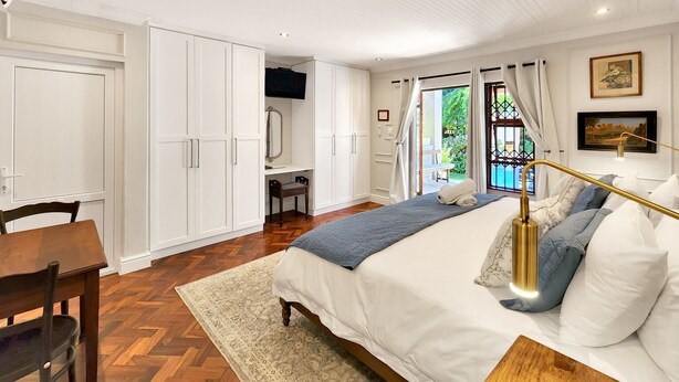 A bedroom with a bed, a desk, and a chair. There is a window with curtains, a door, and a painting on the wall. - Clivia - Pinoak Self-catering Cottages (Cape Town)