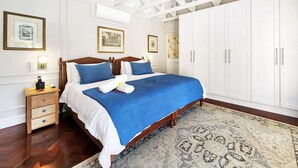 A hotel room with a large bed, wooden headboard, white walls, and a ceiling with exposed beams. There are framed pictures on the wall, a nightstand with a lamp, and a wardrobe with silver handles. - Petunia - Pinoak Self-catering (Cape Town)