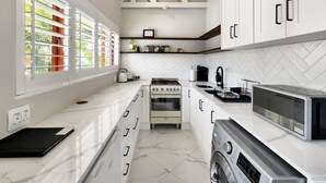 A modern kitchen with white cabinetry, a built-in oven, a microwave, and a large window with blinds. - Petunia - Pinoak Self-catering (Cape Town)