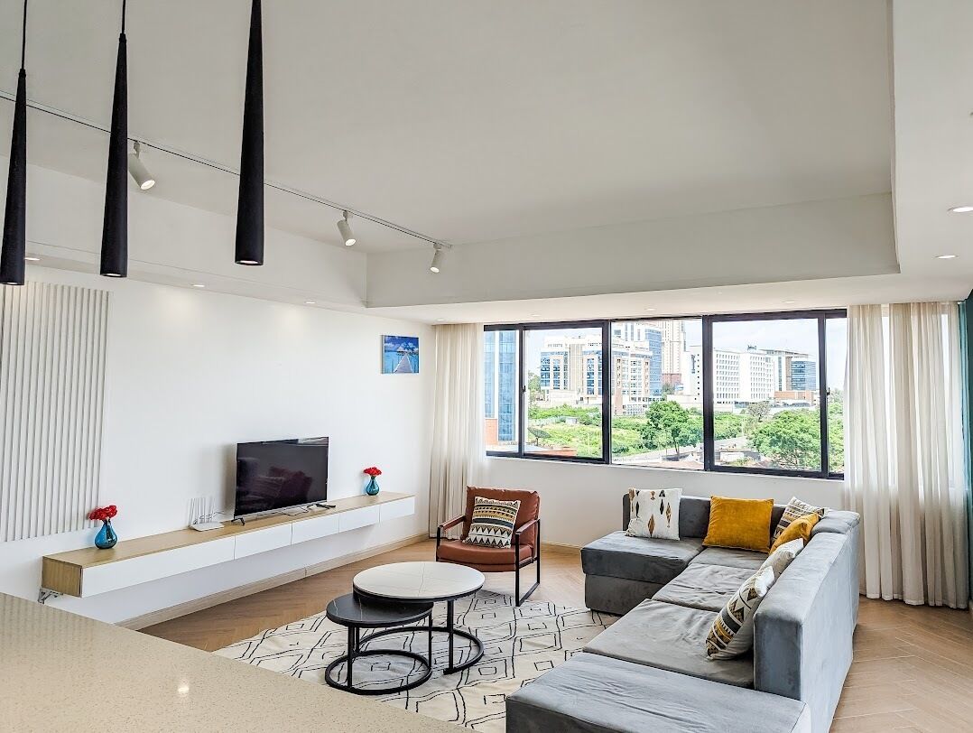 A modern living room with a sofa, a coffee table, and a TV on a white console. Large windows offer a view of buildings and greenery.