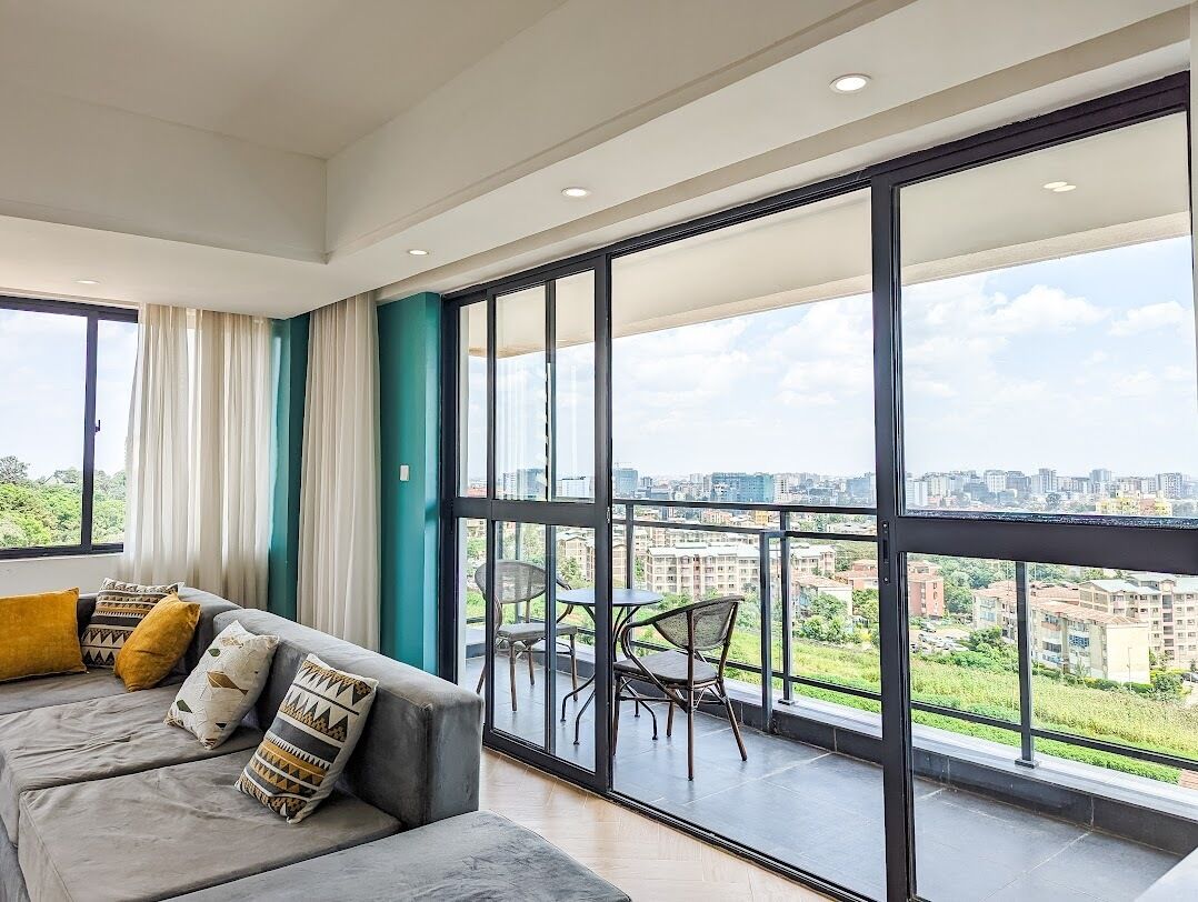 A modern living room with a grey sofa, patterned cushions, and a view of a cityscape through large windows.