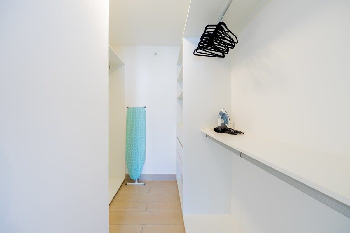 A narrow hallway with a teal ironing board, a black hanger rack, and a small shelf with a telephone.