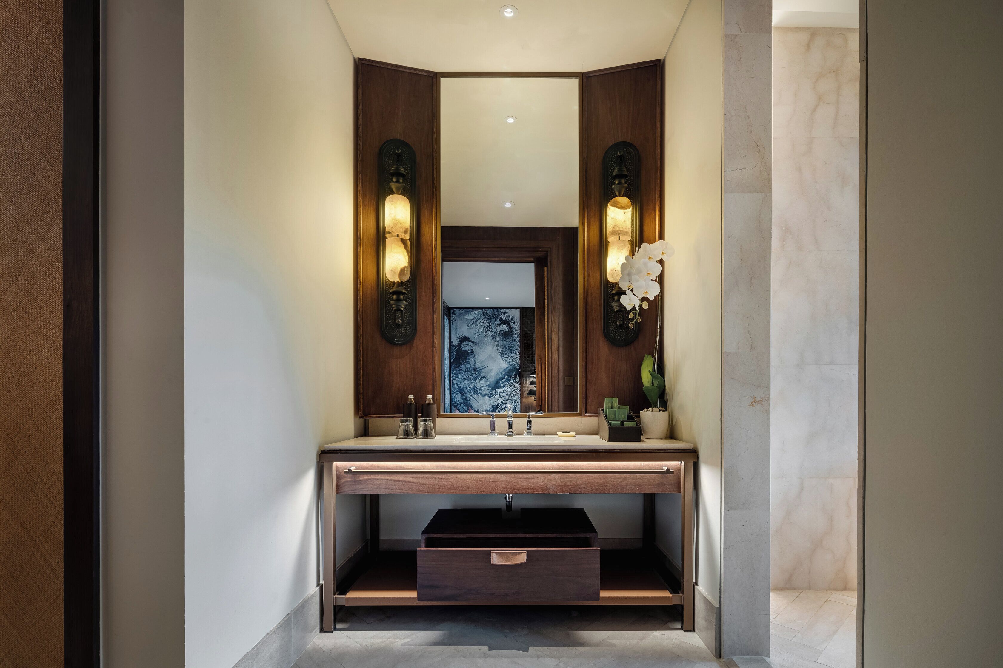 Une salle de bain moderne avec un meuble-lavabo en bois, un miroir agrémenté d’appliques murales et un vase contenant des fleurs blanches.