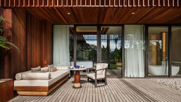 An outdoor living area with a wooden deck, a sofa, a chair, a table, and a large window offering a view of the outdoors. - The Meru Sanur (Denpasar)