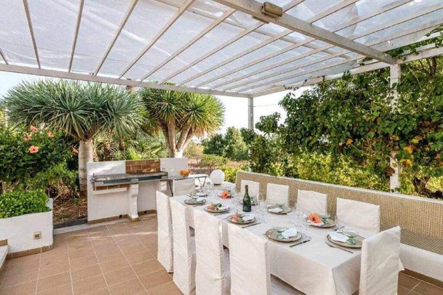 Une terrasse extérieure dotée d’un espace repas couvert, d’un barbecue et d’une végétation luxuriante.