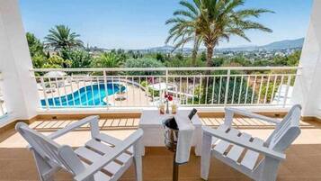 Un balcón con sillas y mesa blancas, con vista a una piscina y palmeras.