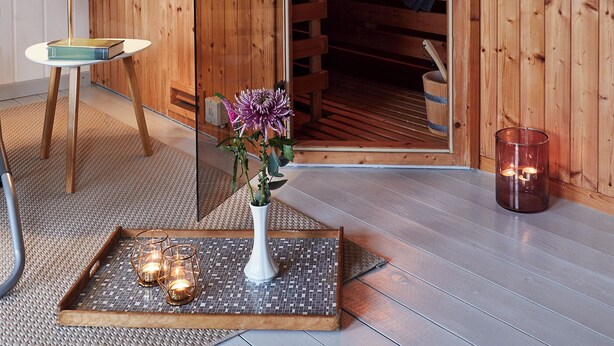 A wooden sauna room with a bench, a towel rack, a small table with a lamp, and a tray with candles.