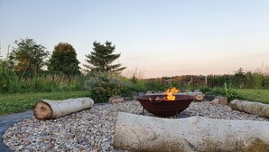 A circular fire pit with logs, surrounded by pebbles and set in a garden with trees and a clear sky.