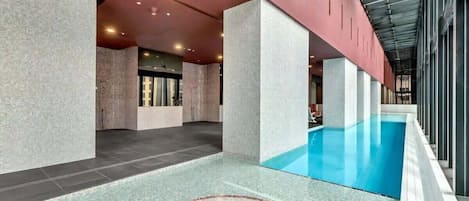 A modern indoor swimming pool with a clear water feature, surrounded by a tiled floor and walls with a textured finish.