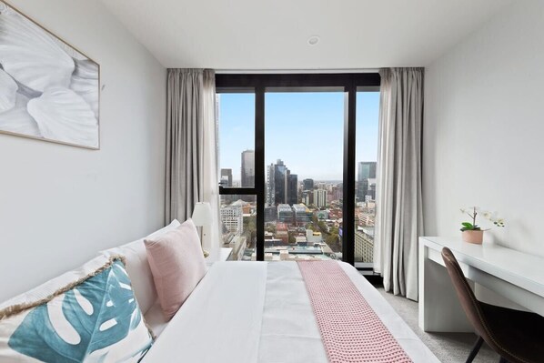 A modern hotel room with a large bed, a desk, and a view of a cityscape through floor-to-ceiling windows.
