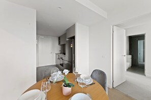A modern dining area with a wooden table set for four, white plates, and wine glasses. A small pink vase with white flowers adds a touch of color. The room has white walls and a visible kitchen area with a refrigerator and cabinets.