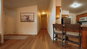 Een hal die leidt naar een keuken met houten kastjes en een magnetron. Er staan twee stoelen in de hal.