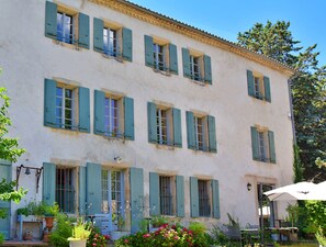 Un bâtiment à deux étages, doté de nombreuses fenêtres et volets, entouré d’une végétation luxuriante et agrémenté d’un espace de détente extérieur.