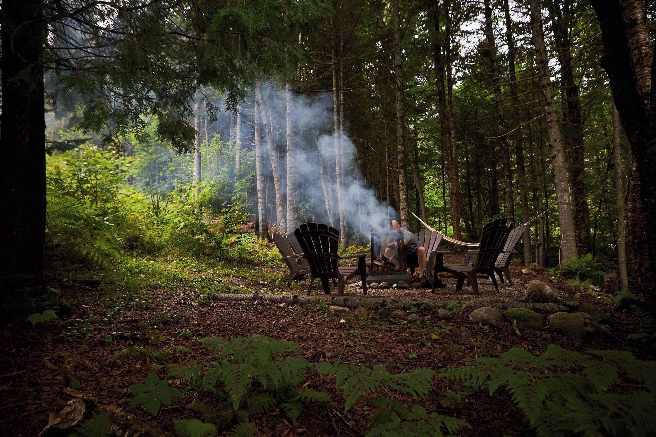 Een boslandschap met twee Adirondack-stoelen, een vuurkorf en rook die opstijgt.