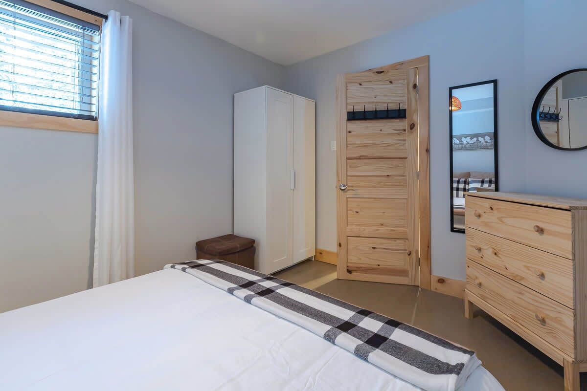 Ein Schlafzimmer mit Bett, Kleiderschrank, einem kleinen Hocker, einer Holztür, einem Spiegel und einem Fenster mit Jalousien.