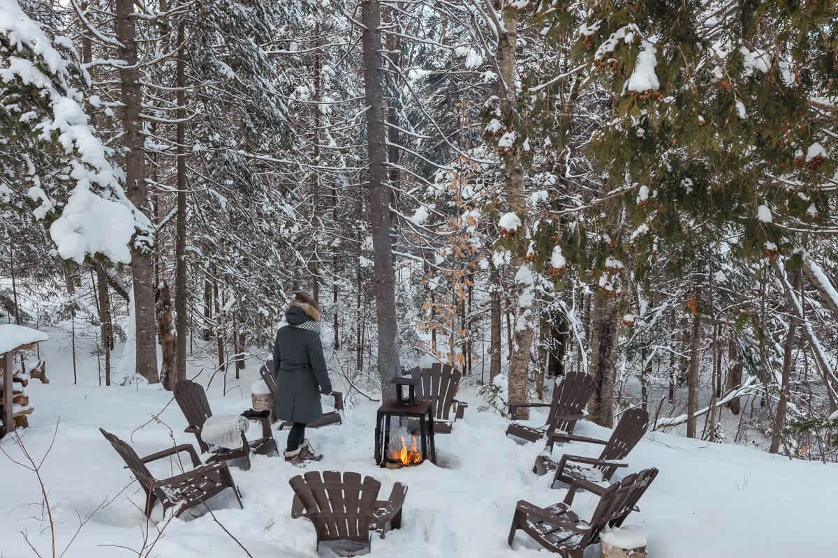A snow-covered forest with wooden chairs arranged around a central fire pit.