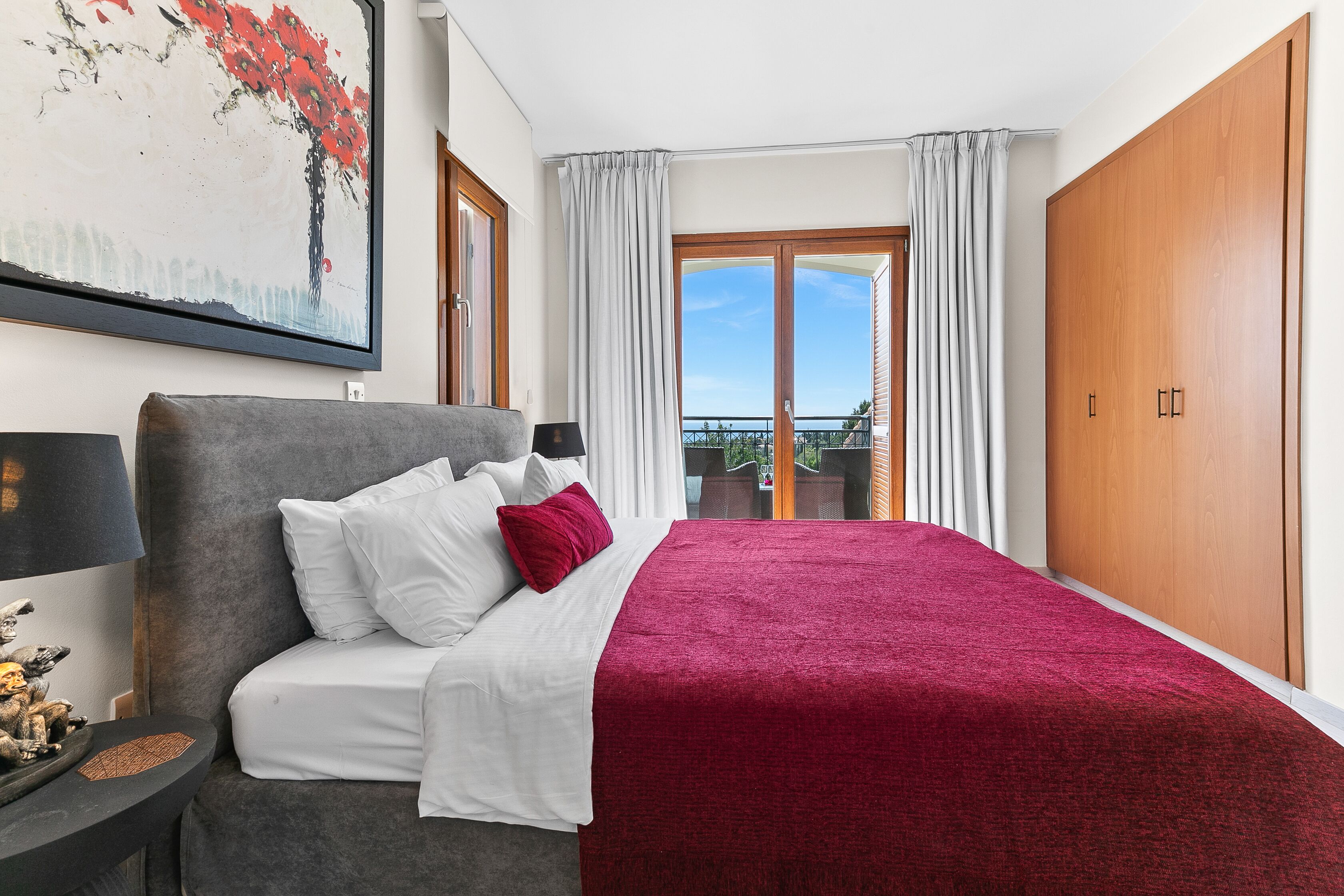A neatly arranged bedroom with a bed, a nightstand, a painting, and a view of the outdoors.