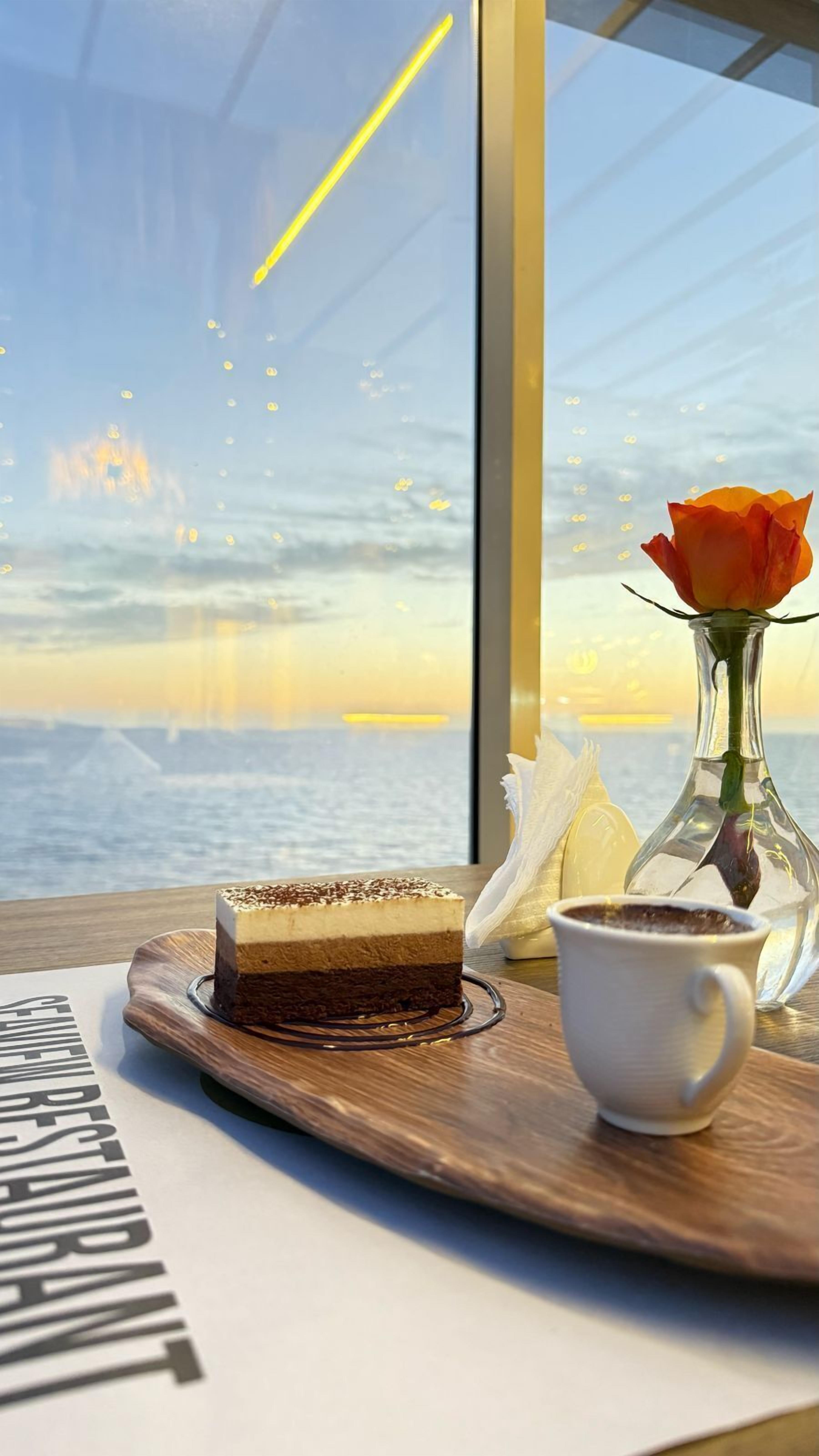 A dessert tray with a slice of cake and a cup of coffee on a wooden table near a window overlooking a body of water.