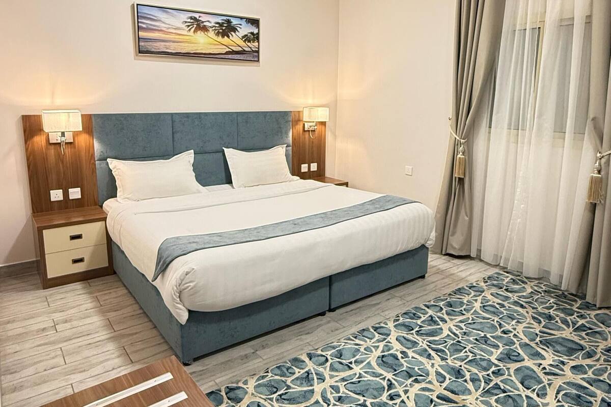 A modern hotel room with a bed, nightstands, a desk, a patterned rug, and a painting on the wall.