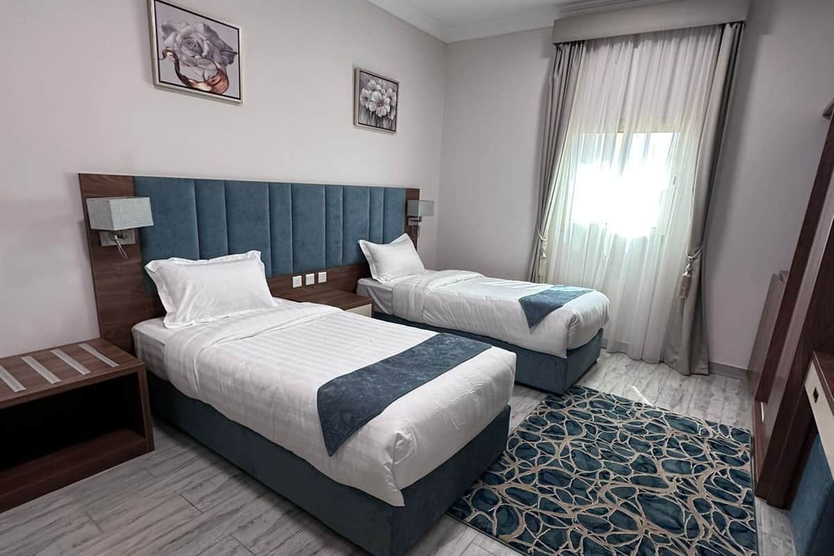 A hotel room with two beds, a wooden nightstand, a blue headboard, a patterned rug, and a window with curtains.