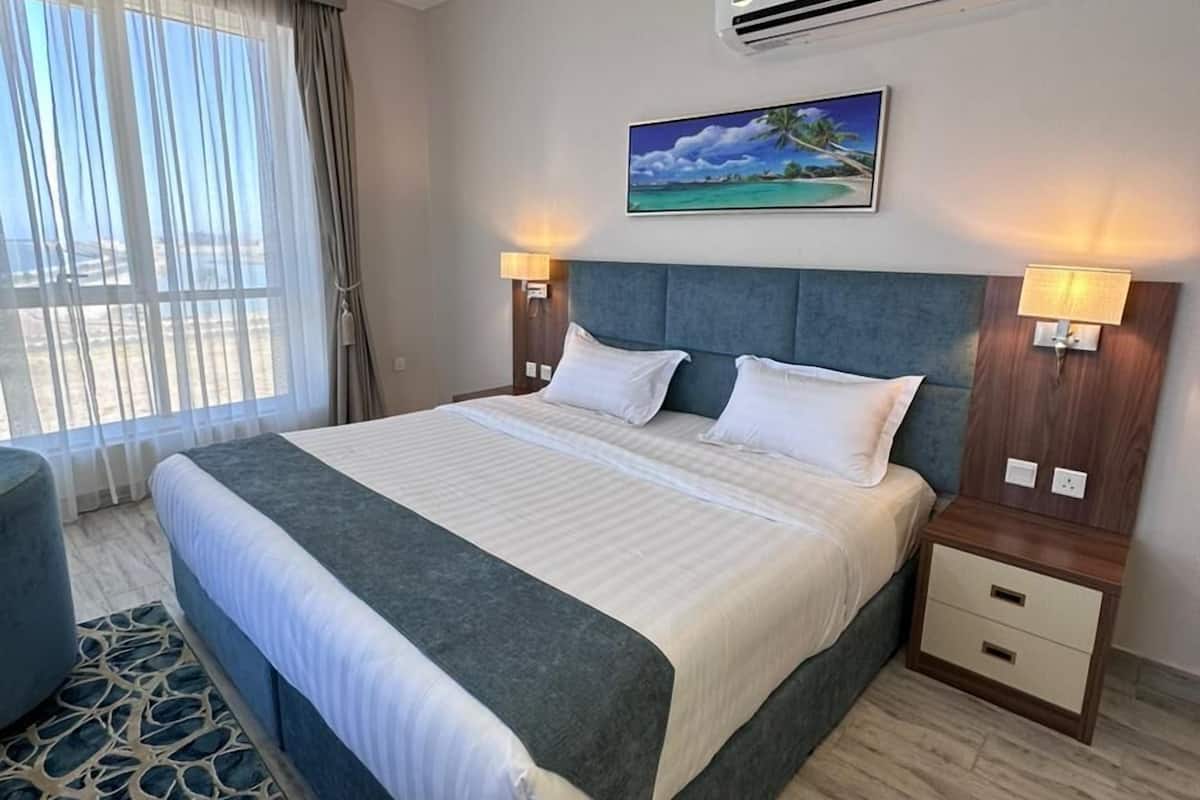 A hotel room with a large bed, a nightstand, a lamp, an air conditioning unit, and a painting of a beach scene.