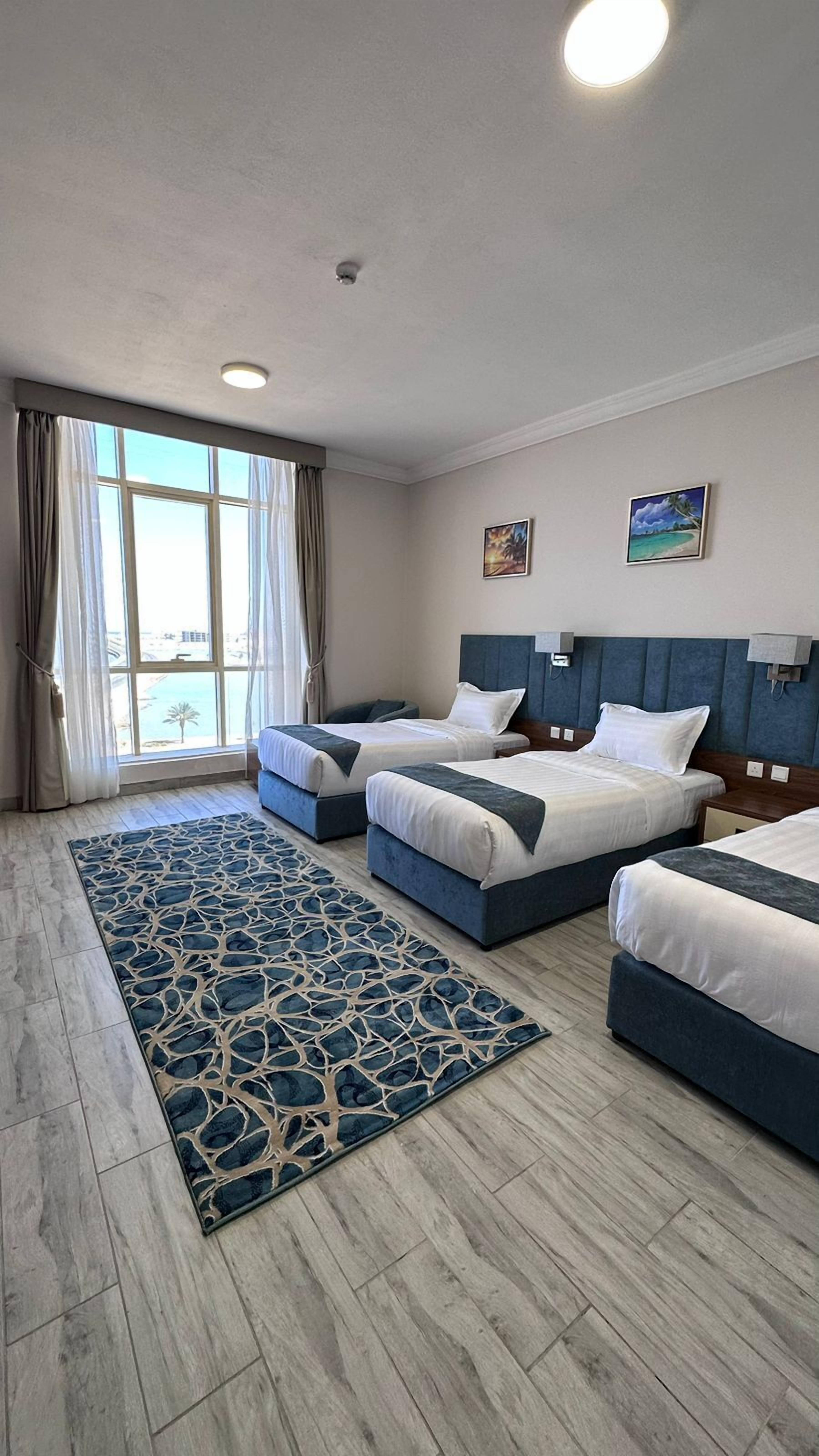 A hotel room with two beds, a large window with curtains, a patterned rug, and artwork on the walls.