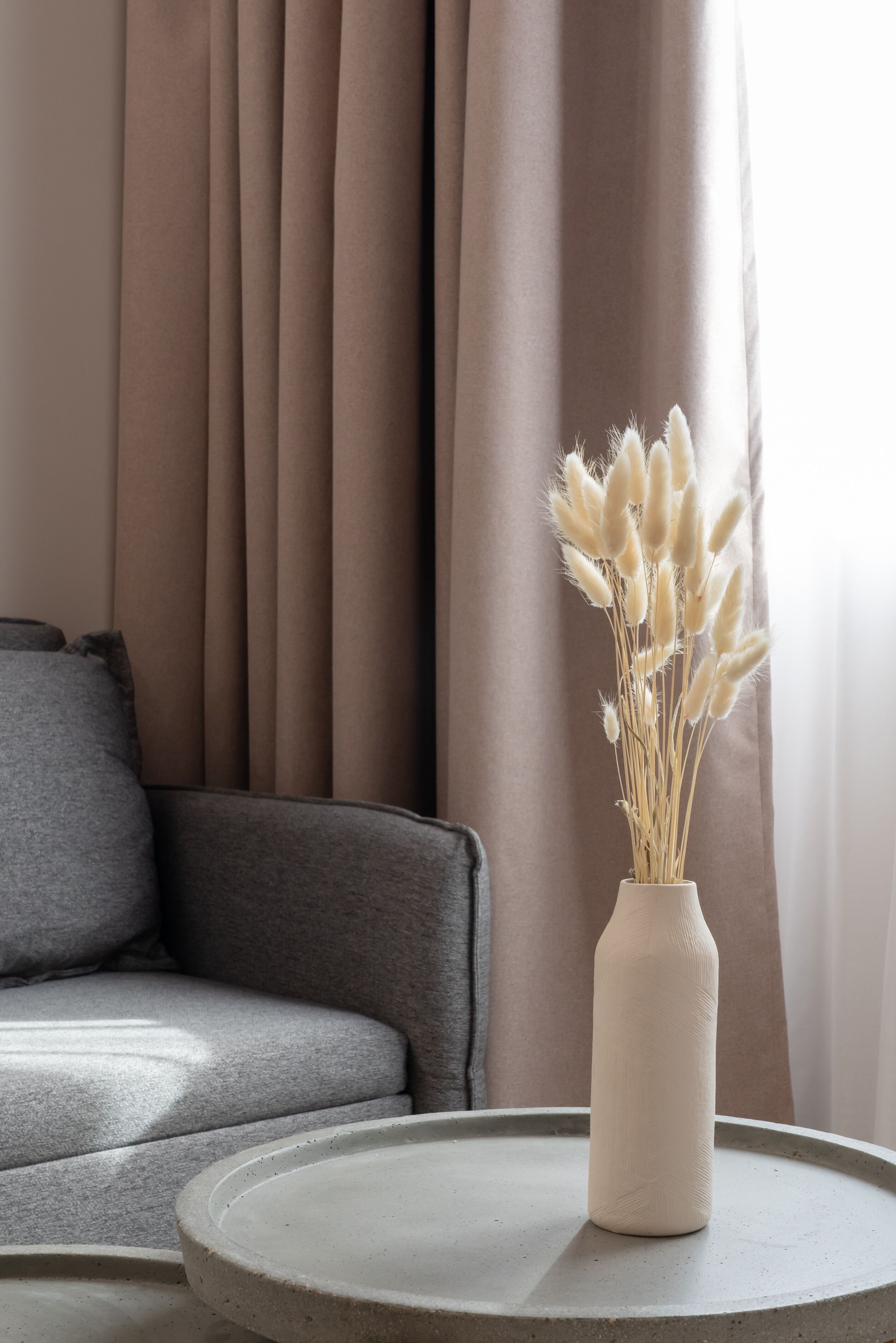 En beige soffa med en kudde, en vit vas med torkat pampasgräs och ett runt bord med en vit tallrik.