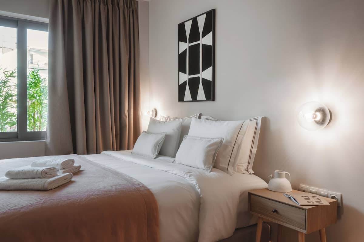 A neatly made bed with white linens and a brown throw blanket, a bedside table with a lamp, and a wall-mounted abstract art piece.