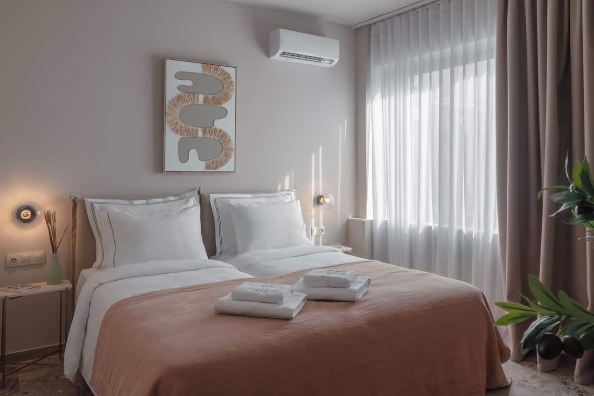 A modern hotel room with a large bed, a bedside table with a lamp, a wall-mounted air conditioner, and a large window with curtains.