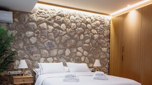 A bedroom with a stone wall, a bed with white linens, wooden flooring, and a nightstand with a lamp. - Num Suites & Lofts Athens (Athens)