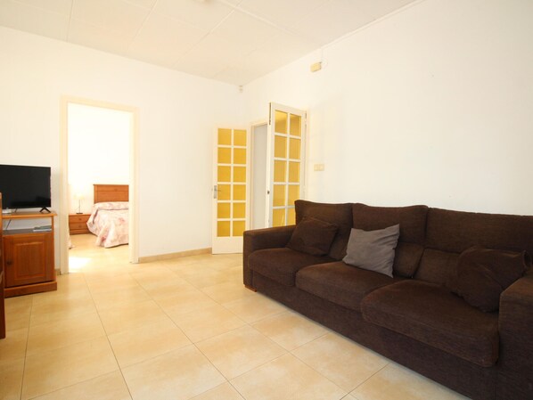 A living room with a brown sofa, a TV on a wooden stand, and a bed with a pink blanket.