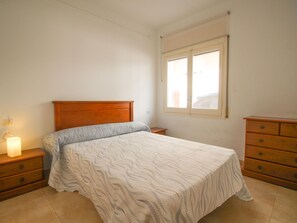 Ein Schlafzimmer mit hölzernem Kopfteil, einem Bett mit gemustertem Bettbezug, einem Nachttisch mit Lampe, einer Kommode und einem Fenster mit Jalousien.