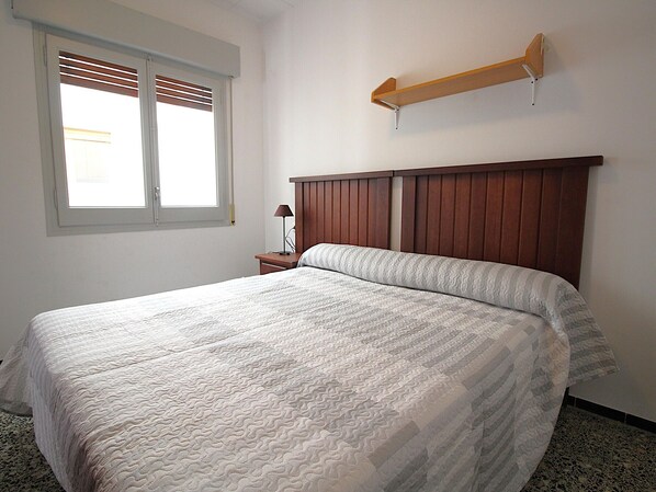 Ein Schlafzimmer mit einem Bett, einem Nachttisch, einer Lampe, einem Fenster mit Jalousien und einem Holzregal.