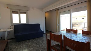A room with a blue sofa, a dining table with chairs, a small white table, and a window with blinds. - Penthouse avec Grande Terrasse, à 100 m de la Plage de Llançà, Rénové, avec WIFI en Été (Llançà)
