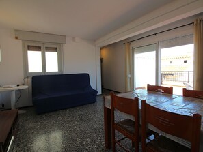A room with a blue sofa, a dining table with chairs, a small white table, and a window with blinds. - Penthouse avec Grande Terrasse, à 100 m de la Plage de Llançà, Rénové, avec WIFI en Été (Llançà)