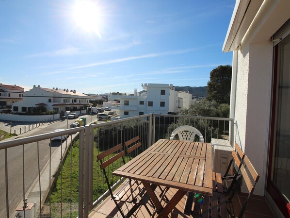 Un balcon avec une table et des chaises en bois, donnant sur une rue bordée de voitures garées et d’immeubles.