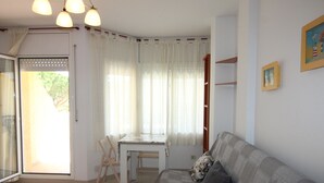 Ein Wohnzimmer mit grauem Sofa, einem kleinen weißen Tisch und einem Esstisch mit Stühlen. Eine Glastür führt auf einen Balkon.