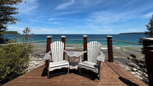 Zwei Adirondack-Stühle auf einer Holzterrasse mit Blick auf ein Gewässer unter blauem Himmel.