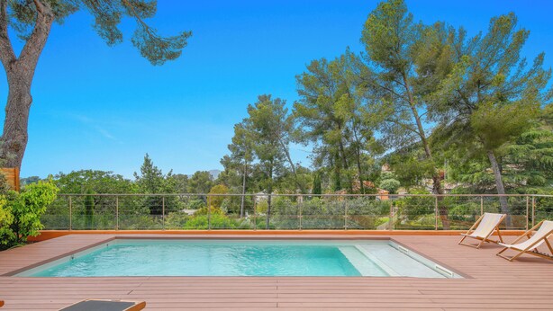 A rectangular pool with a wooden deck, surrounded by a metal fence, and overlooking a landscape with trees and buildings. - Californie-piscine/CLiM/WiFi 12P (Carqueiranne)