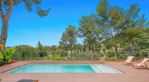 A rectangular pool with a wooden deck, surrounded by a metal fence, and overlooking a landscape with trees and buildings. - Californie-piscine/CLiM/WiFi 12P (Carqueiranne)