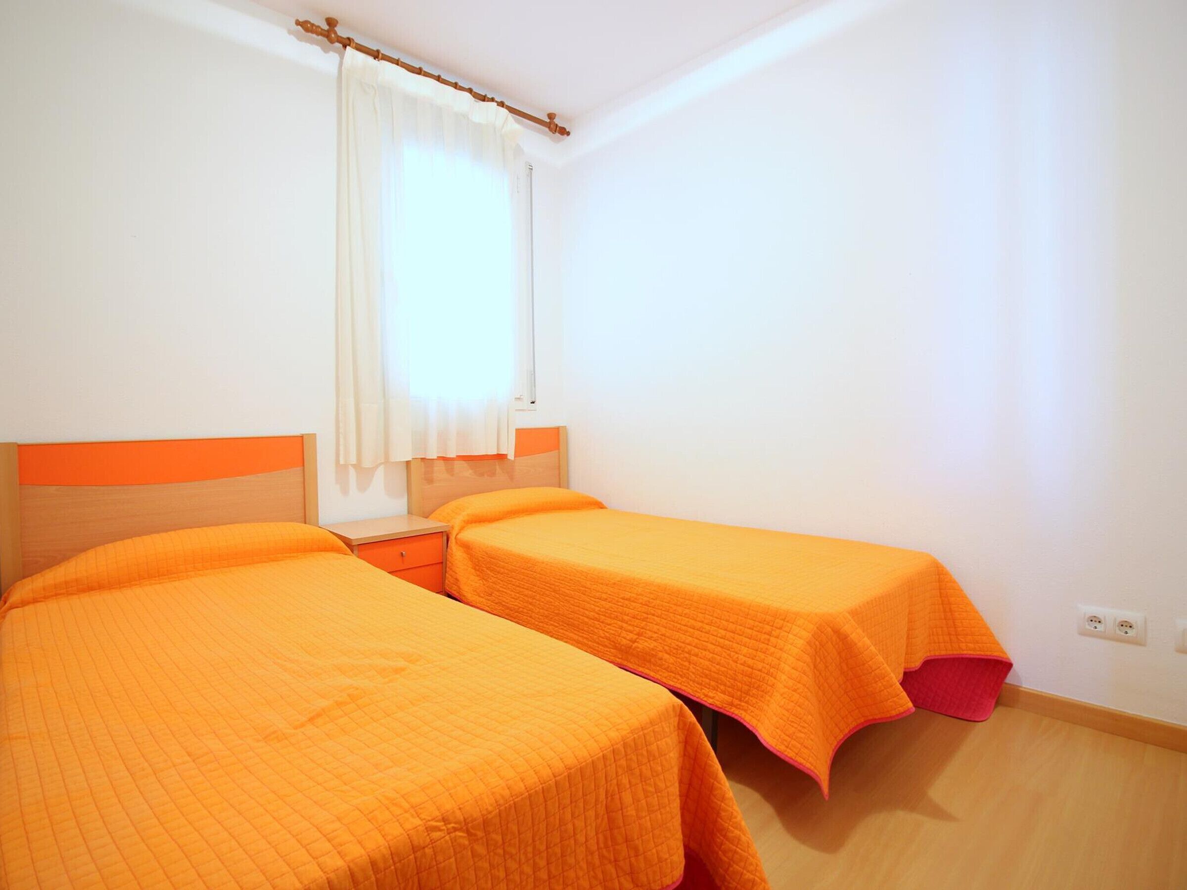 A room with two beds, each with an orange bedspread, a wooden headboard, and a window with a white curtain.