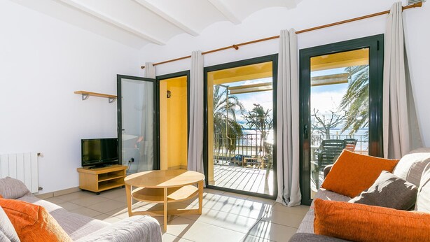 Una sala de estar moderna con sofá, televisor y una pequeña mesa. Amplias puertas correderas de vidrio dan a un balcón con vistas a palmeras y al mar.