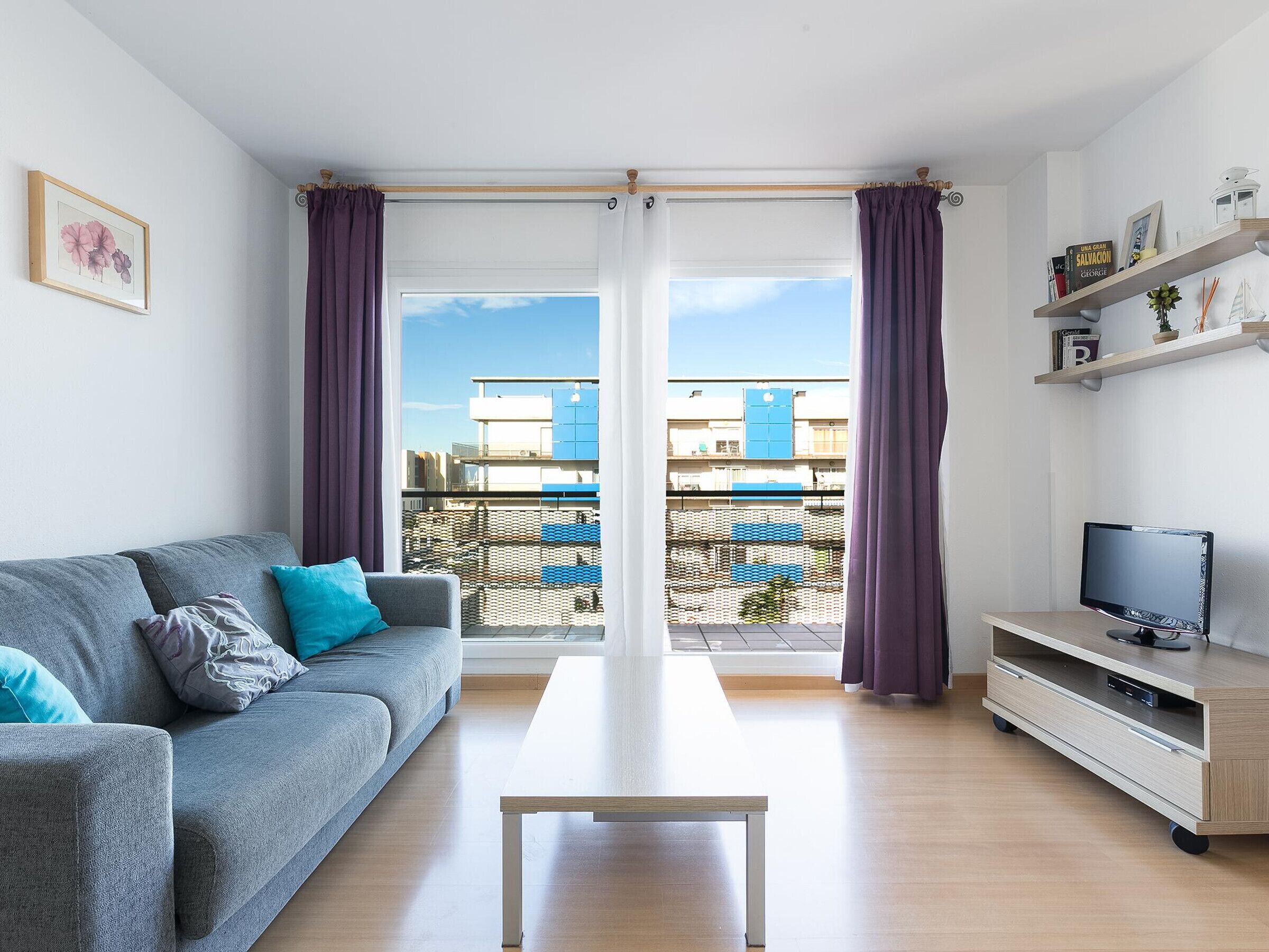 A modern living room with a grey sofa, a wooden coffee table, a TV on a wooden stand, and a balcony with purple curtains.