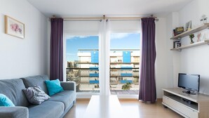 Una sala de estar moderna con un sofá gris, una mesa de centro de madera, un televisor sobre un mueble de madera y un balcón con cortinas moradas.